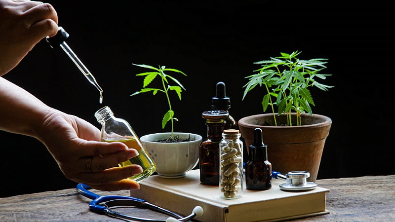 a hand taking CBD oil from a bottler with CBD capsules and tinctures on a wooden tray with cannabis plant on pots and stethoscope