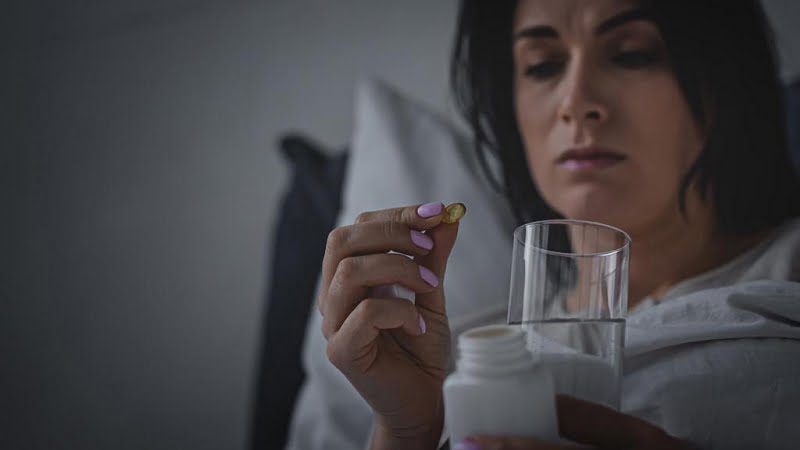 woman with sleep disorder holding glass of water and capsule in bed