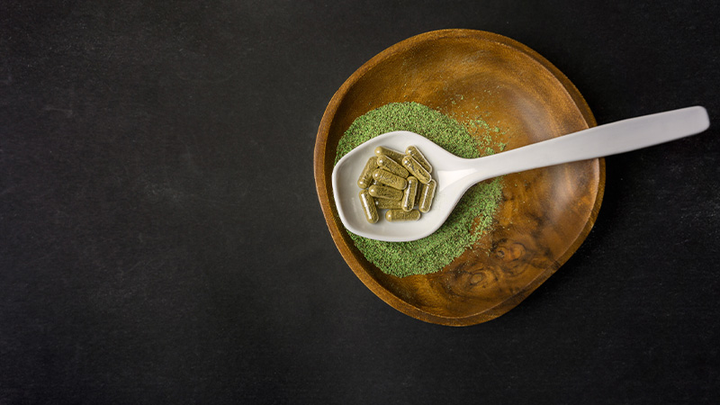 Kratom powder in a wooden bowl with a spoon of Kratom capsules
