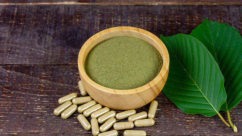 Kratom powder in a wooden bowl and some Kratom capsules on the table.