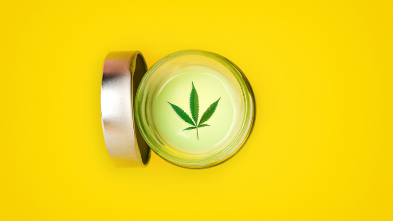 A jar of CBD cream with hemp leaf on top on a yellow background